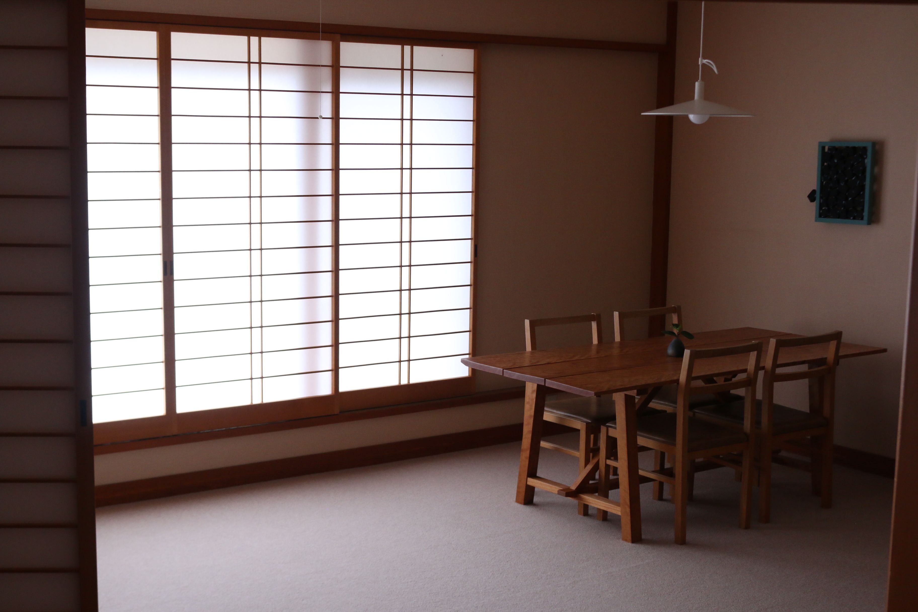 A traditional Japanese room with tatami flooring, a sliding shoji screen, a wooden dining table with chairs, and a hanging light fixture.