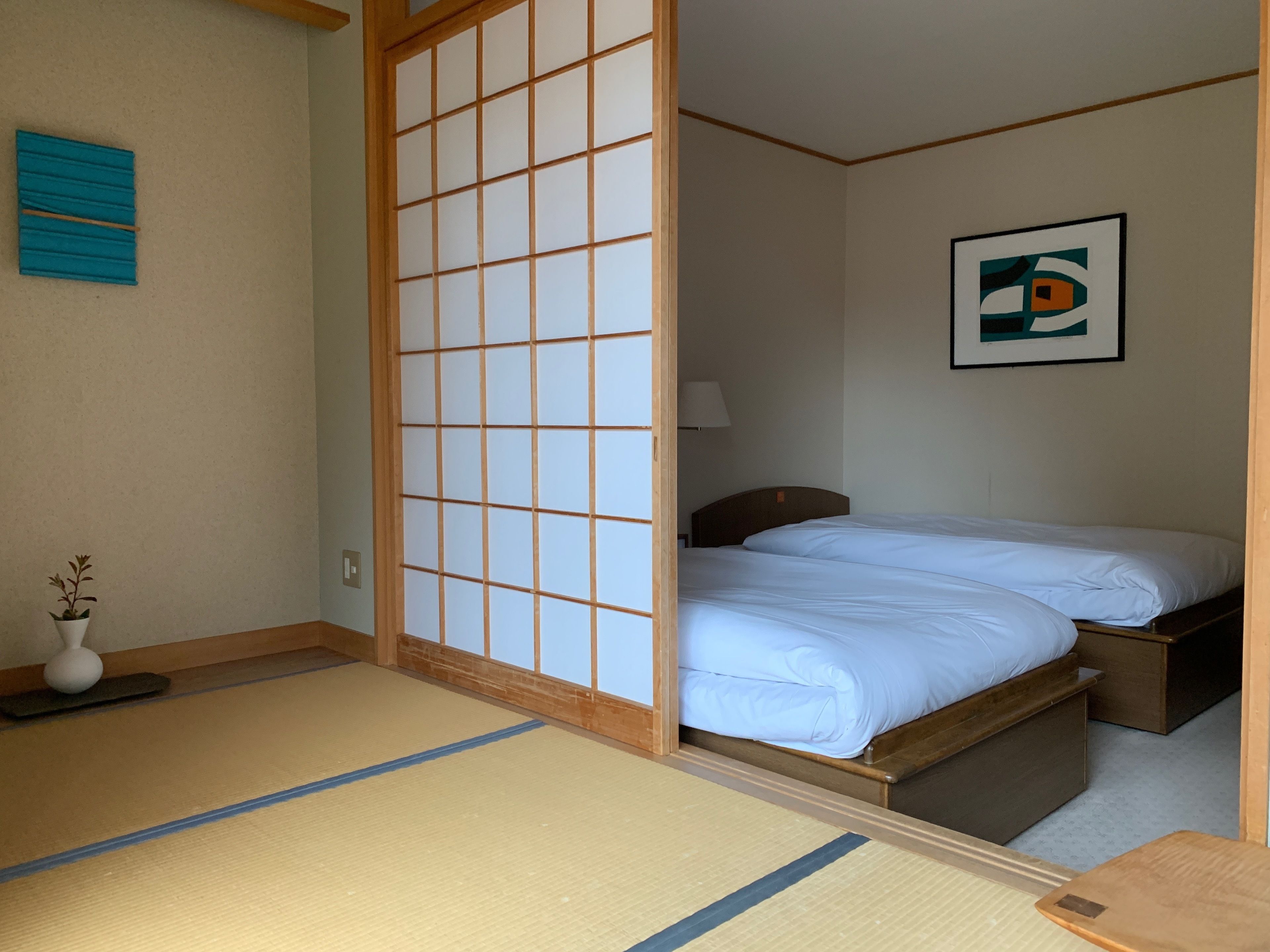Une pièce de style japonais traditionnel, avec un sol recouvert de tatamis, une porte coulissante en bois, un lit avec une literie blanche, une petite table de chevet, une lampe et un tableau encadré accroché au mur.
