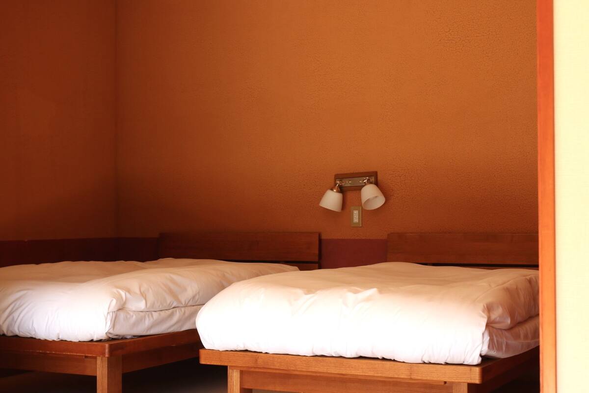 Dos camas de madera con ropa blanca en una habitación con paredes naranja y una lámpara fijada a la pared.