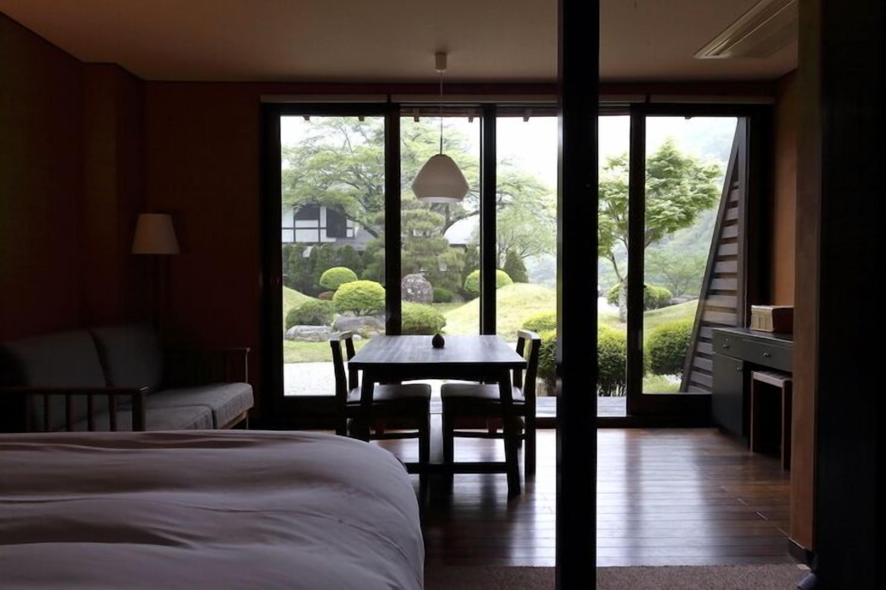 Quarto com cama, escrivaninha e poltrona, sofá e vista para um jardim através de amplas janelas.