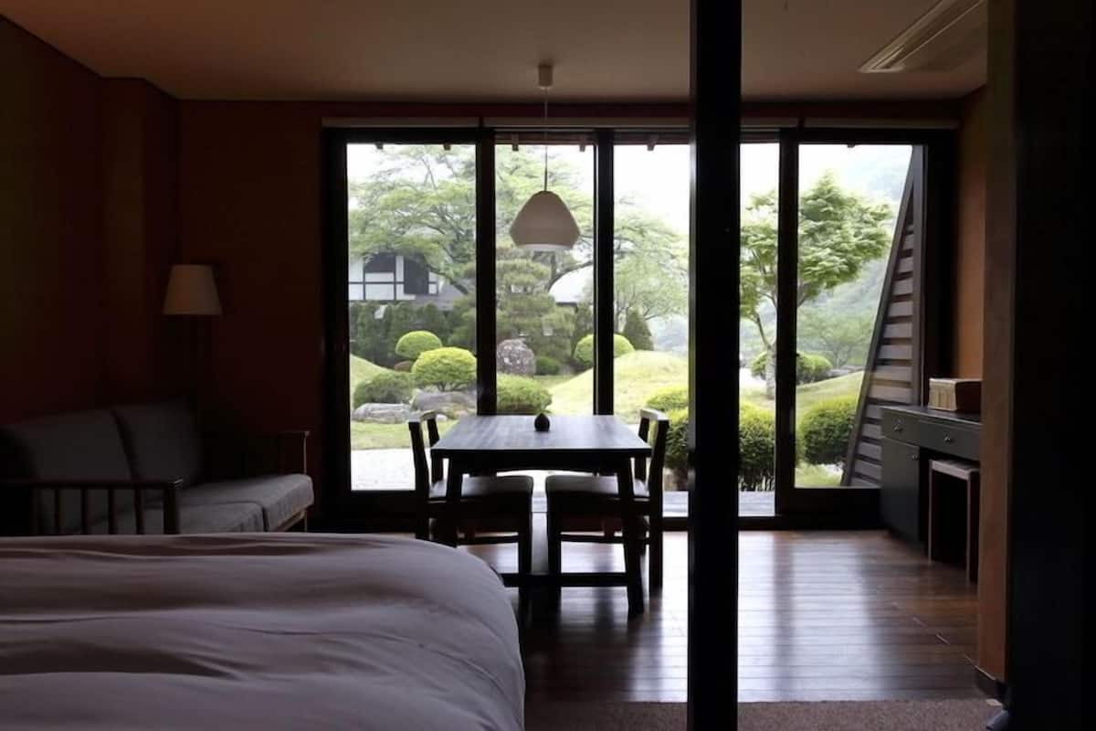 Quarto com cama, escrivaninha e poltrona, sofá e vista para um jardim através de amplas janelas.