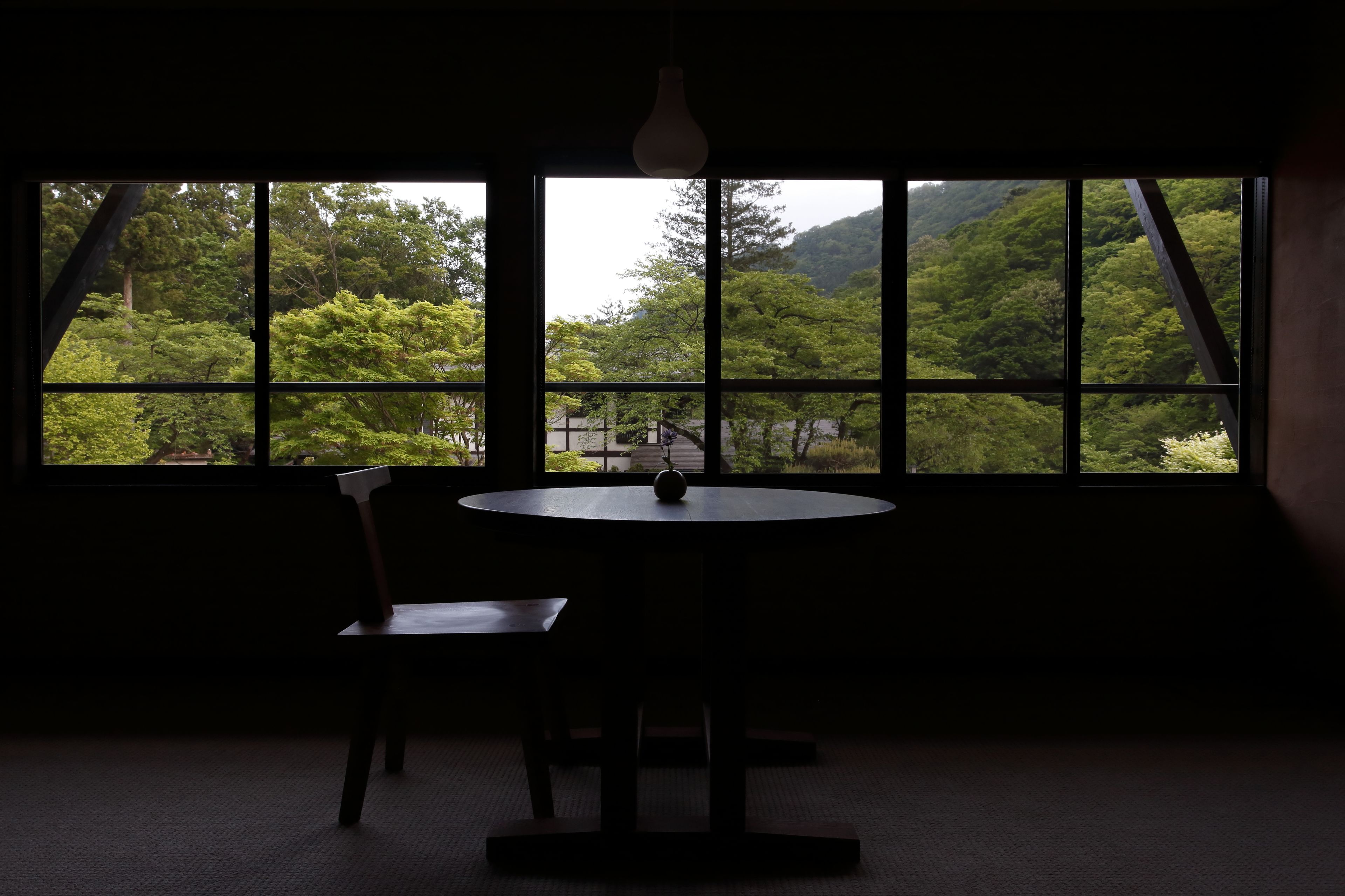 Une pièce avec une table ronde et une chaise, un luminaire suspendu et une grande fenêtre donnant sur une zone boisée.