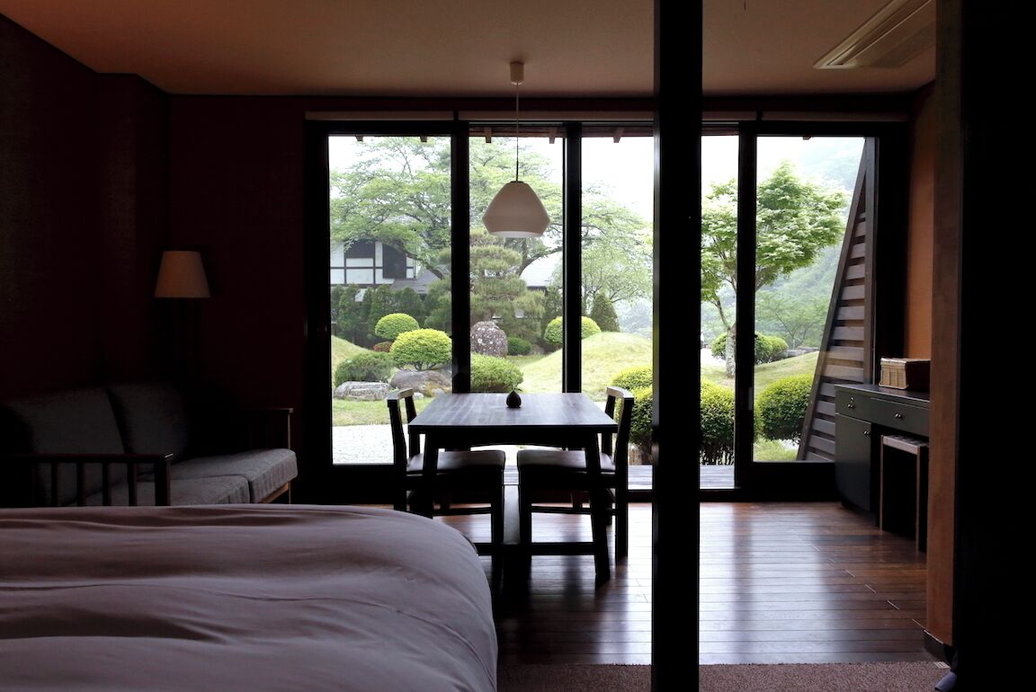 Ein Zimmer mit Bett, Tisch, Stühlen, einer Lampe und Blick auf einen Garten durch große Fenster.