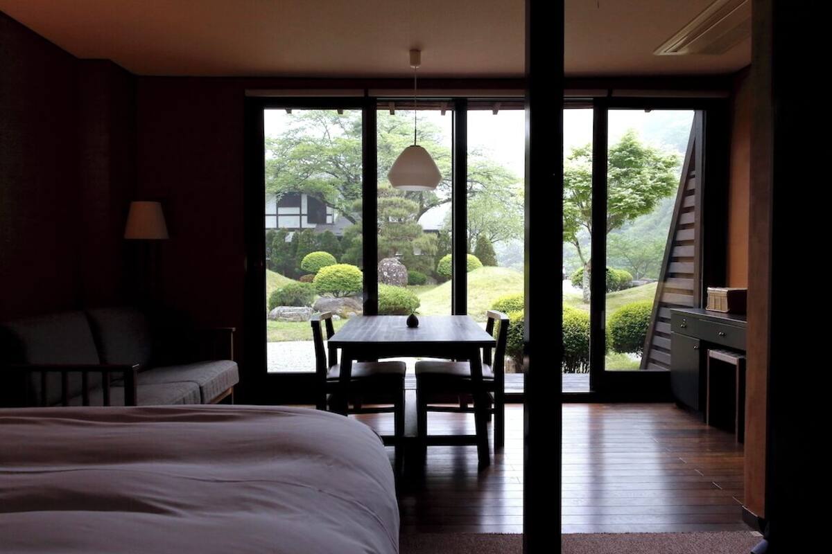 Una habitación con cama, una mesa y sillas, una lámpara y vistas a un jardín a través de amplios ventanales.