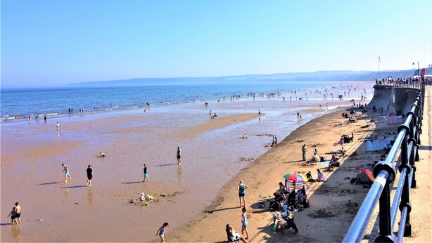 A beach with a concrete barrier, people walking, and umbrellas. - ❤️ Large Family Self Catering Holiday House-Filey (Filey)