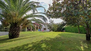 Ein gepflegter Garten mit einer Palme, einer Rasenfläche und einem Gebäude mit rotem Dach und Bögen.