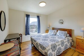 Un dormitorio con cama, escritorio, silla, ventana con cortinas, mesita de noche con lámpara y una planta en la pared.