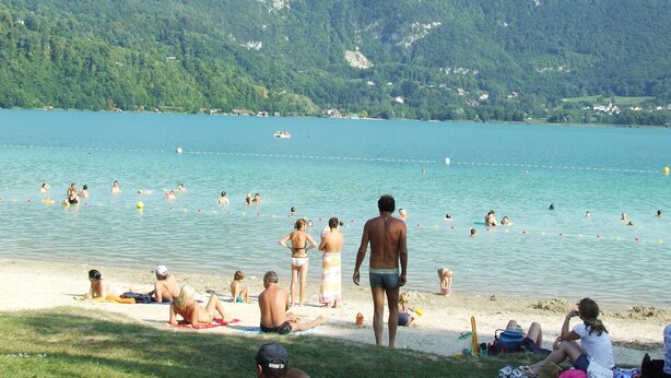 Ein Strand am See mit badenden und sonnenden Menschen, umgeben von Bergen.