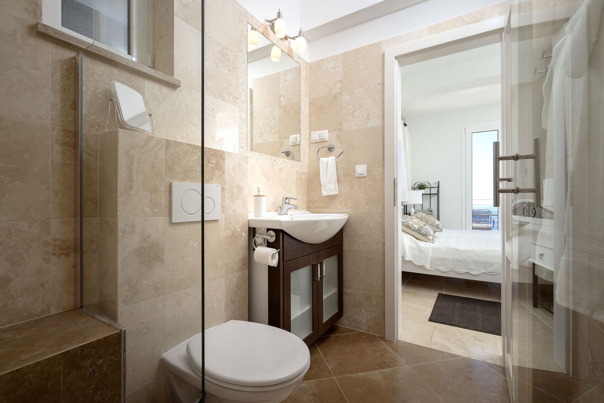 A modern bathroom with a glass shower enclosure, a white toilet, a sink with a mirror, and a bed visible through an open door.