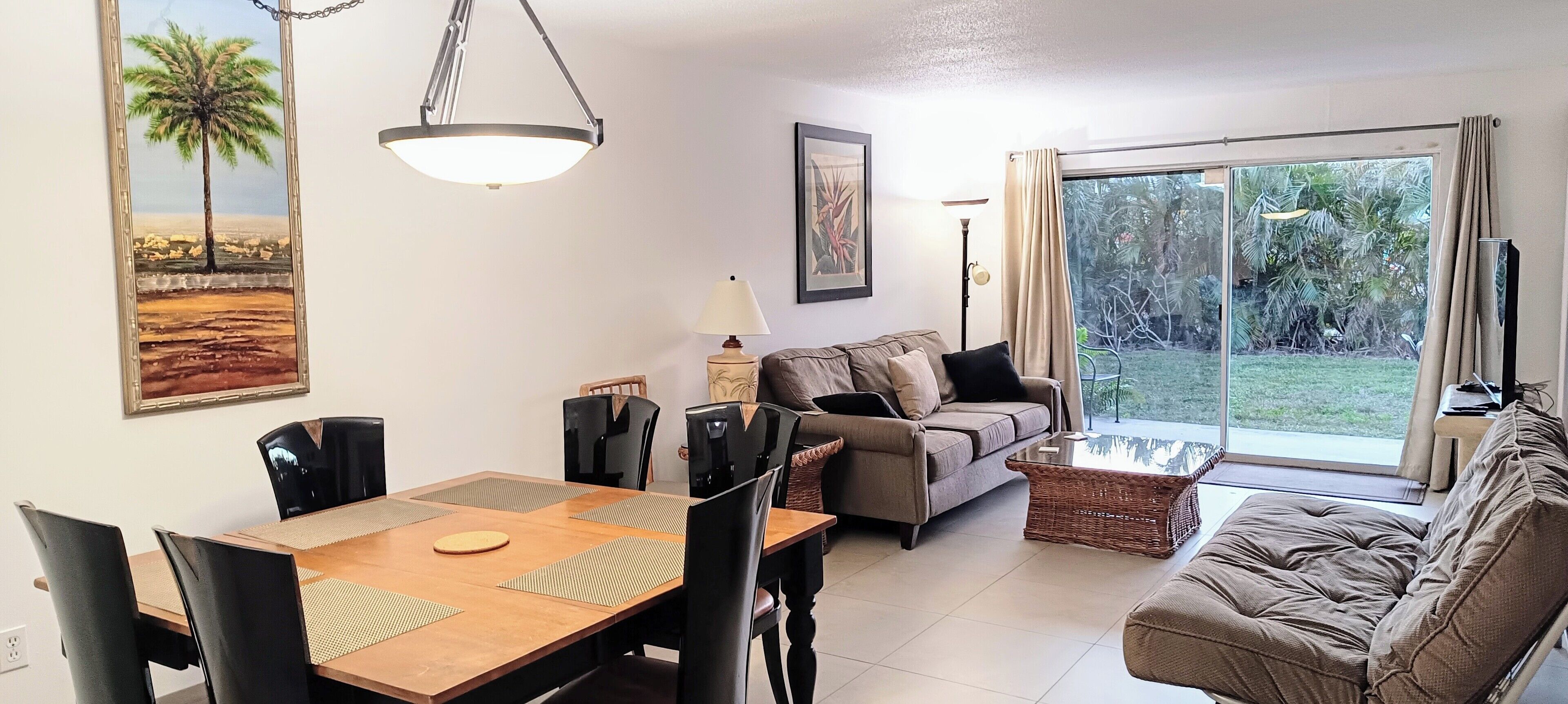 Een woonkamer met een bank, een eettafel met stoelen, een lamp en een groot raam met uitzicht op groen.