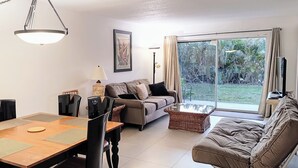 A living room with a sofa, a dining table with chairs, a lamp, and a large window with a view of greenery. - Near Siesta Key. Lg Patio. 2 King beds, Wi-Fi, Updated Sarasota Condo. 1st floor (Sarasota)