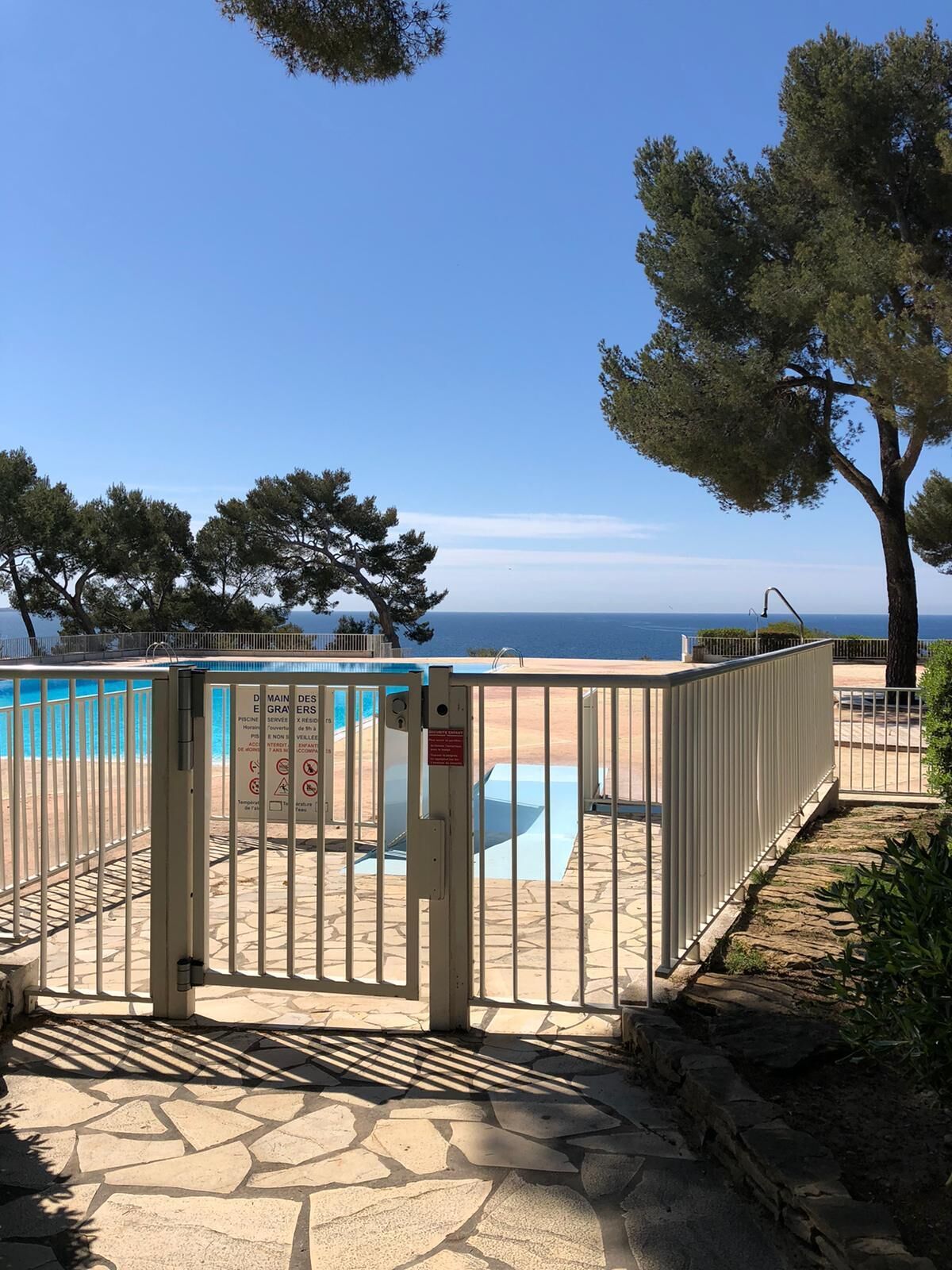 A gated pool area with a clear view of the sea, surrounded by stone flooring and trees.