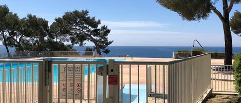 A gated pool area with a clear view of the sea, surrounded by stone flooring and trees.