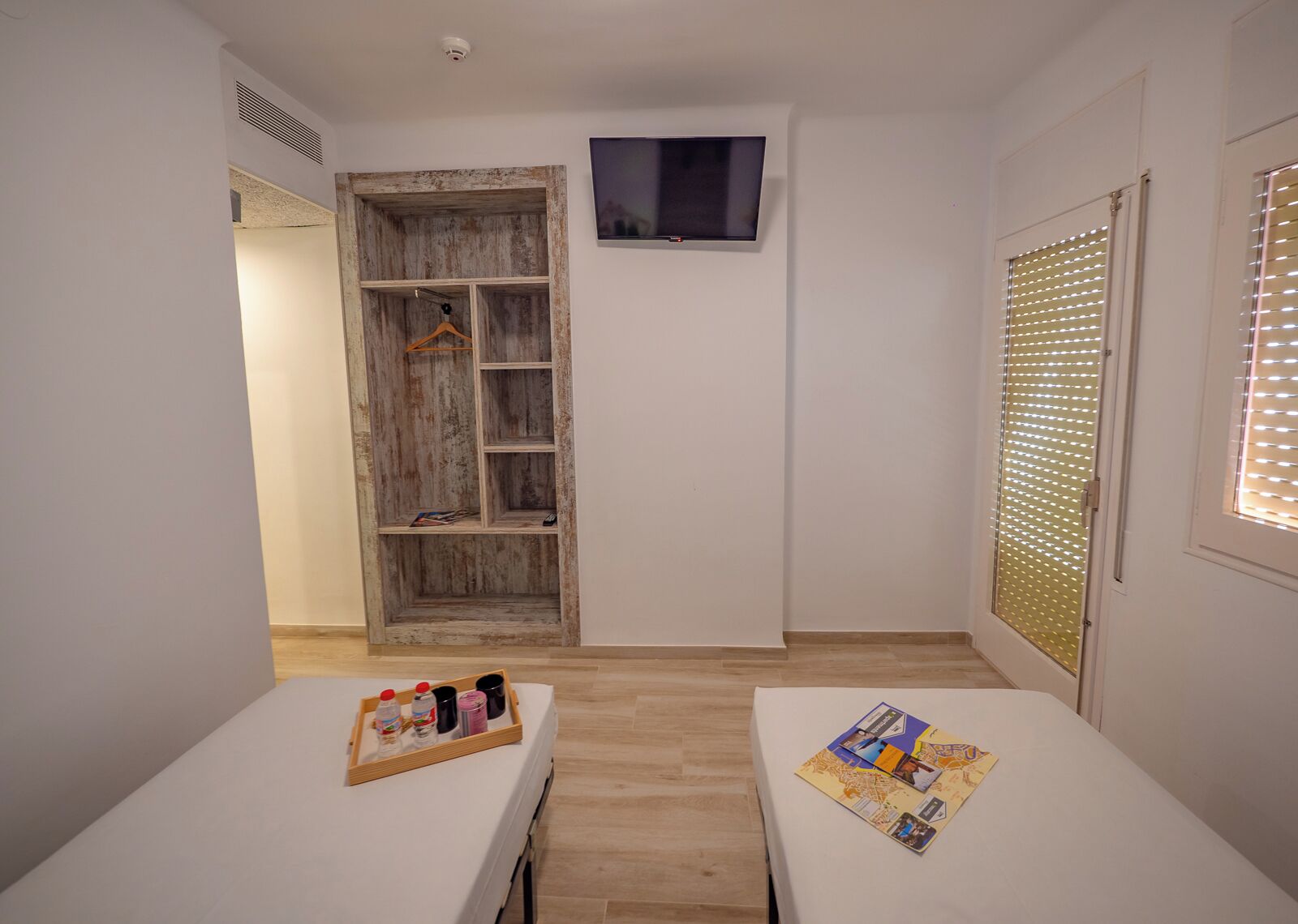 A room with a wooden wardrobe, a television mounted on the wall, and a window with blinds.