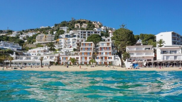 Un pueblo costero con edificios en una ladera, una playa de arena y aguas azules cristalinas.