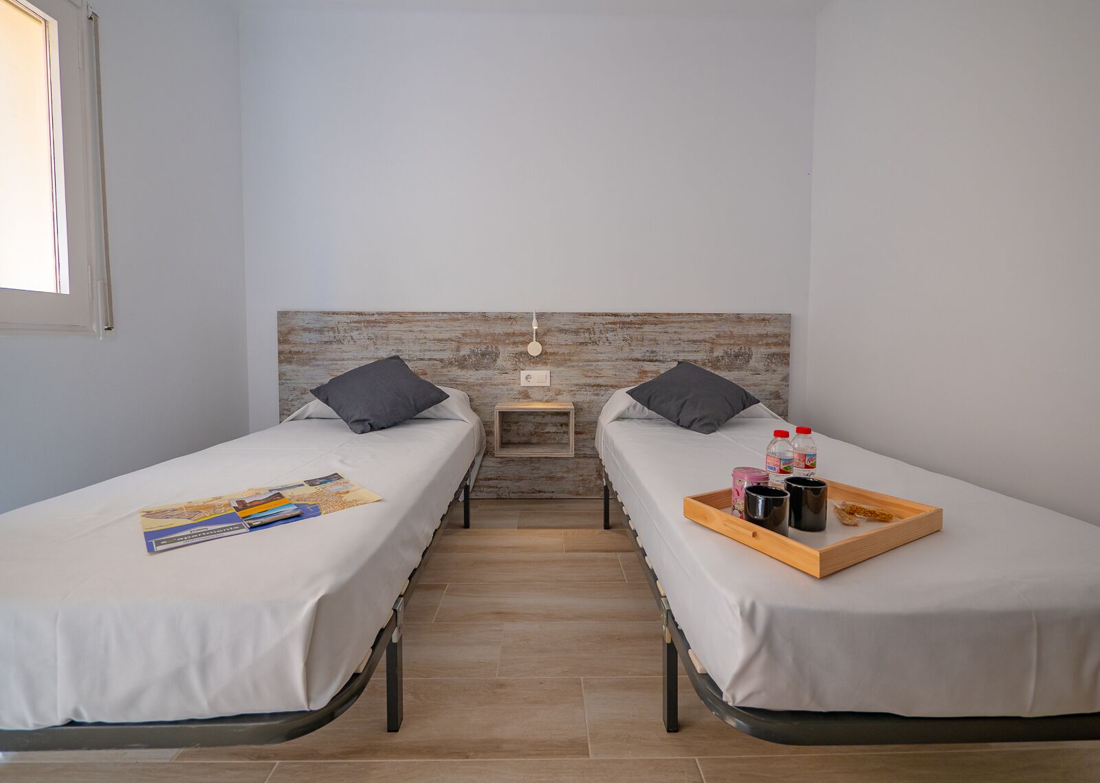 Une petite chambre propre, avec deux lits simples, une tête de lit en bois, une petite table de chevet et un plateau contenant des articles de toilette.