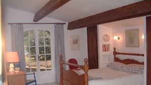 A bedroom with a wooden bed, a nightstand, a lamp, a chair, and a window with curtains. - Idyllic St Tropez location with pool. Tranquil setting close to all the action. (Ramatuelle)