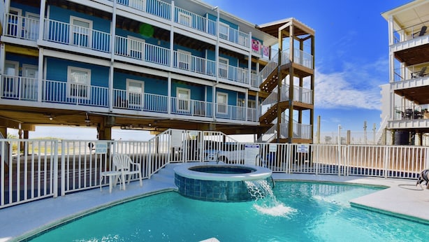 A swimming pool with a hot tub, surrounded by a white fence, and a multi-story building with balconies in the background. - Recently refurnished condo directly on the beach! (Gulf Shores)