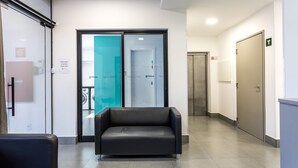 A modern hotel lobby with a black leather sofa, a glass door leading to a service area, and a T-shaped sign indicating the direction to the elevator. - Flat completo com garagem, 2 camas de casal, perto do Centro e Jd Bot, PUC (Rebouças)