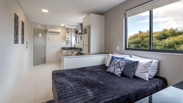 A modern bedroom with a bed, a window with a view of greenery, and a kitchenette in the background. - Flat completo com garagem, 2 camas de casal, perto do Centro e Jd Bot, PUC (Rebouças)