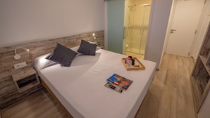 Una cama bien tendida con almohadas, una mesita de noche con una lámpara, una bandeja con botellas y una revista.