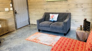 A room with a grey sofa, a red patterned ottoman, a wooden door, and a wall-mounted light fixture. -  Knotty Squirrel Unit 2 King Suite with Ladder Loft (Mountain View)