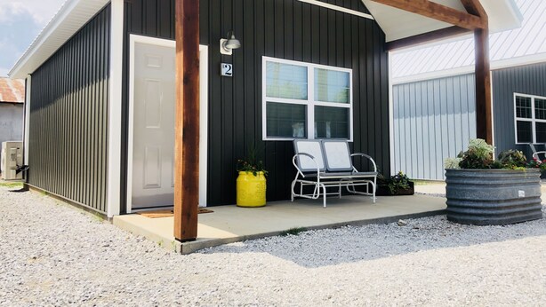 A building with a dark exterior, white trim, and a number '2' sign. There is a covered porch with two chairs and a yellow planter. -  Knotty Squirrel Unit 2 King Suite with Ladder Loft (Mountain View)