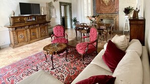 Un salon spacieux avec un plafond haut, un décor orné, un grand téléviseur, un canapé, des fauteuils, une table à manger et un tapis à motifs.