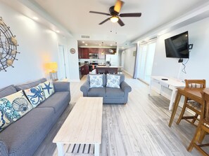 A living room with a sofa, a coffee table, and a TV. There is a kitchen in the background.