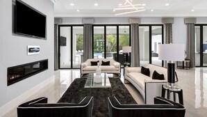 A modern living room with a flat-screen TV, fireplace, and a large window offering a view of the outdoors. - White House at Reunion | 10BR Luxe Stay w/ Bowling Alley, Arcade, Private Pool, and Gym Near Disney (Kissimmee)
