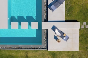 Vista aérea de uma piscina com um deck revestido, cadeiras de descanso e uma mesa com uma bebida.