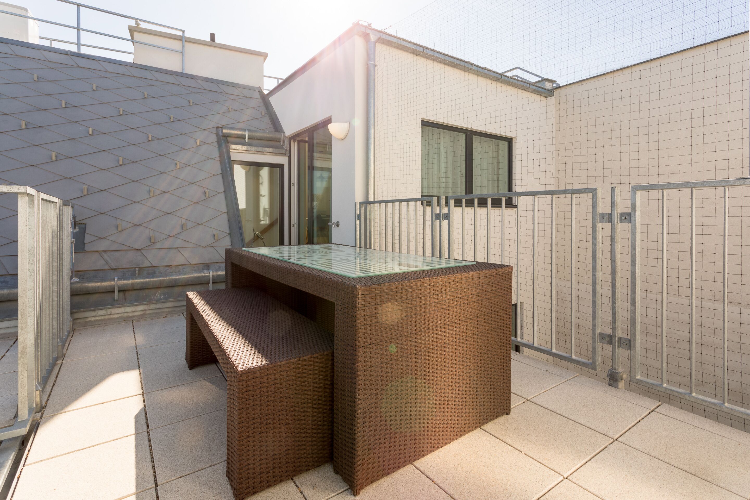 A rooftop terrace with a glass-top table and wicker furniture, enclosed by metal railings and a tiled floor.