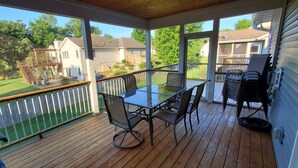 A covered outdoor deck with a glass table and chairs, a barbecue grill, and a view of a backyard with a wooden deck and a lawn.