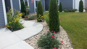 Ein Wohnhaus mit gepflegtem Vorgarten, der einen Betonweg, einen kleinen Garten mit gelben Blumen und ordentlich geschnittenen immergrünen Bäumen zeigt.