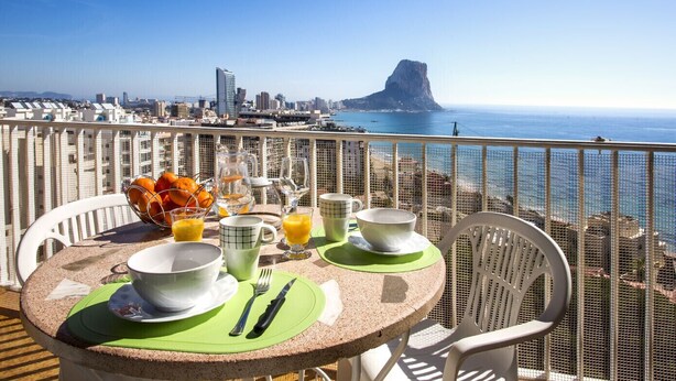 Une table à manger dressée avec des aliments pour le petit-déjeuner, offrant une vue sur l’océan et le paysage urbain.