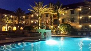 Ein Pool bei Nacht mit Wasserfall, umgeben von Palmen und beleuchteten Gebäuden.