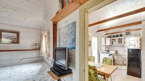 Uma cabana aconchegante com interior de madeira, uma cama com colcha florida, uma televisão em um móvel, uma área de jantar com mesa e cadeiras, e uma cozinha com armários e uma geladeira.
