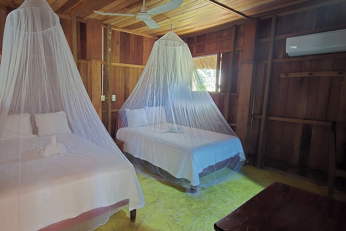 Two beds with mosquito netting, wooden walls, a ceiling fan, and an air conditioning unit.