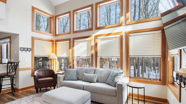 A spacious living room with a large sectional sofa, a small round table, and a patterned rug. The room features multiple large windows with wooden frames and blinds, offering views of trees and a snowy landscape. - Ski In/Ski Out on Boyneland, Slps 14 (Boyne Falls)