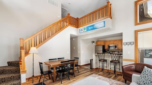 A spacious living area with a wooden staircase, a dining table with chairs, a kitchen area with a microwave, and a sofa with a patterned throw. - Ski In/Ski Out on Boyneland, Slps 14 (Boyne Falls)