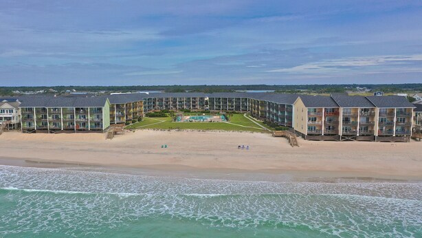 Un hotel frente a la playa con varios balcones, una piscina y una playa al fondo.
