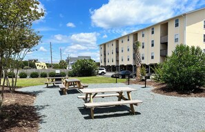 A courtyard with picnic tables and a building in the background. - Surf Condo 518 - HeavenLee By The Sea - First Floor End Unit (Surf City)