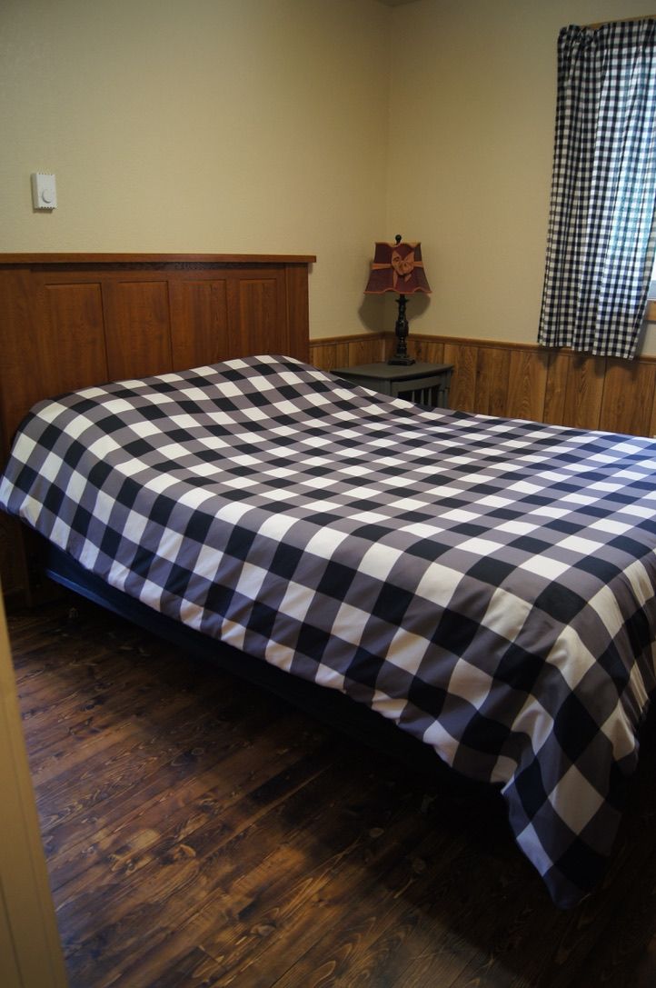 Une chambre à coucher avec une couvre-lit à carreaux, une tête de lit en bois, une petite table avec une lampe et une fenêtre ornée de rideaux à carreaux.