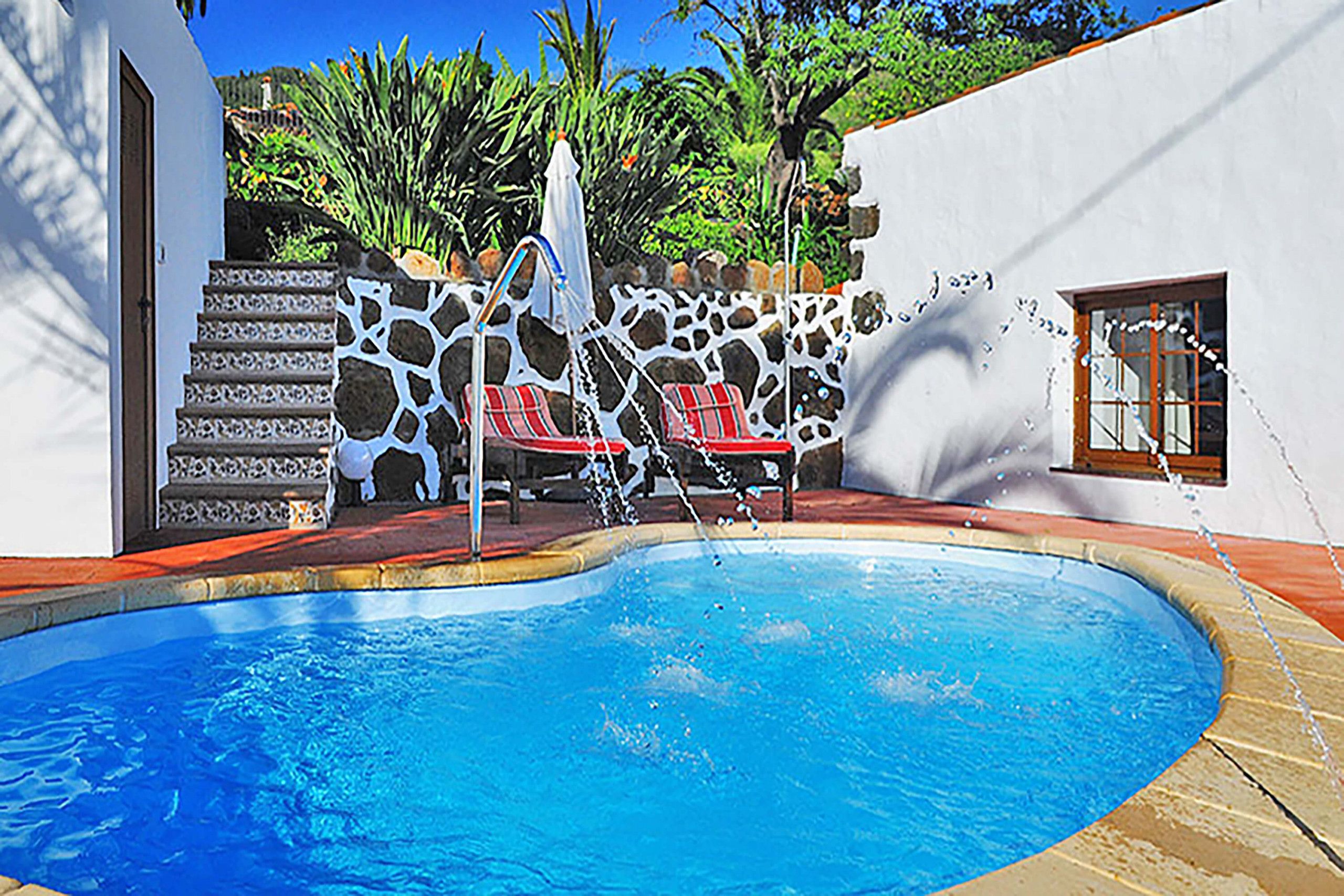 A swimming pool with a stone wall, red lounge chairs, and a white fence.