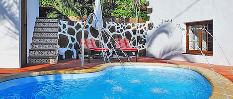 A swimming pool with a stone wall, red lounge chairs, and a white fence.