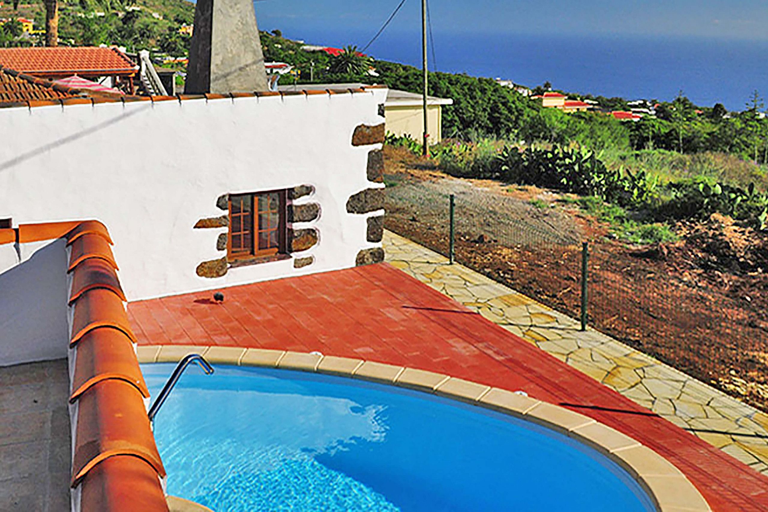 Una piscina con cubierta roja, una pared blanca con ventana y vista al mar y casas al fondo.