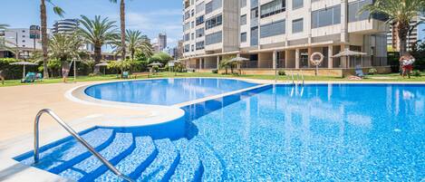 Una piscina de diseño moderno, rodeada de palmeras y un edificio de varios pisos con balcones.
