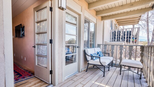 A covered porch with a wooden floor, a sliding glass door, a chair with a cushion, and a small table. - Casa Bohemia | Charming Retreat (Santa Fe)