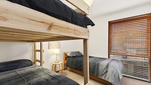 A room with two bunk beds, a small table with a lamp, and a window with blinds. - Ocean's Course (Lorne)