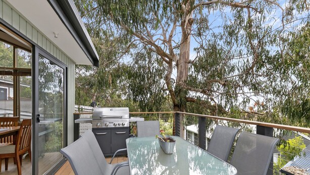 An outdoor deck with a glass table, chairs, and a barbecue grill. - Ocean's Course (Lorne)